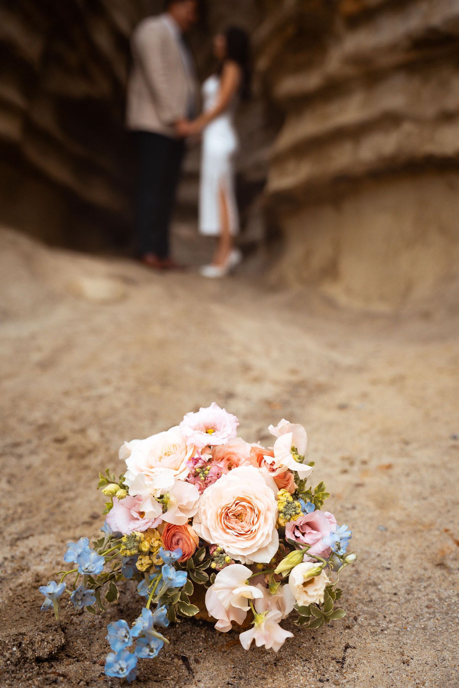 A romantic floral bouquet with pink roses and blue accents against sandy backdrop with couple in background.