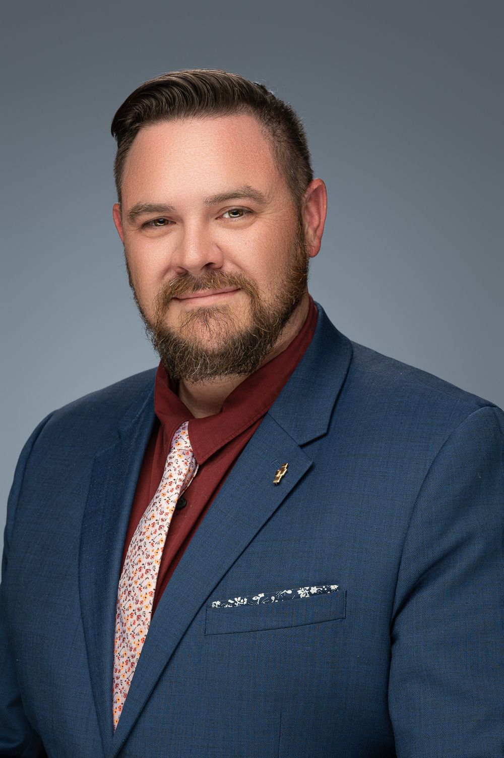 Professional headshot of someone wearing a navy suit jacket with red turtleneck and patterned tie against gray background.