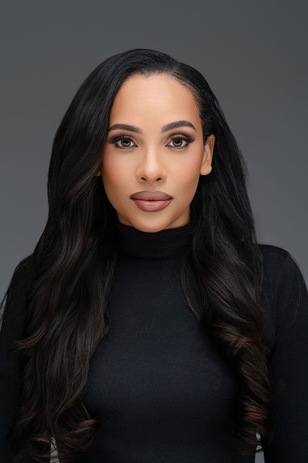 Professional portrait of a woman with long dark wavy hair wearing a black turtleneck against a gray background.