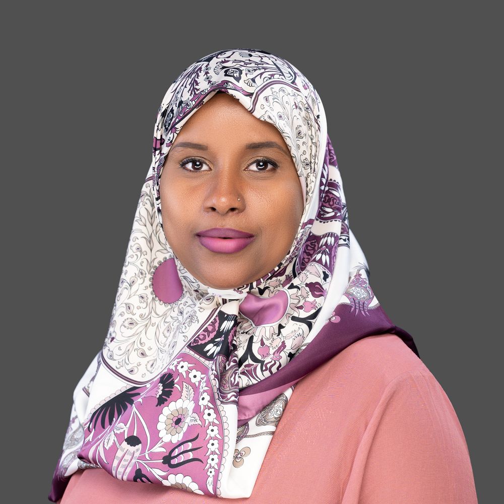 A woman wearing a white and purple patterned hijab and pink lipstick poses against a gray background.
