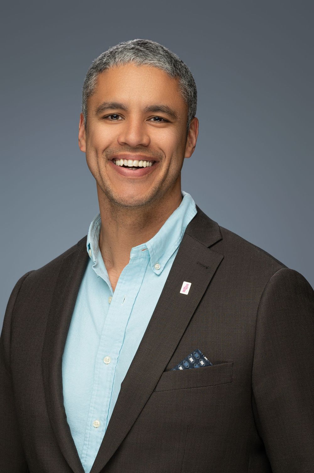 Professional headshot of someone wearing a dark suit jacket and light blue dress shirt with a polka dot pocket square.