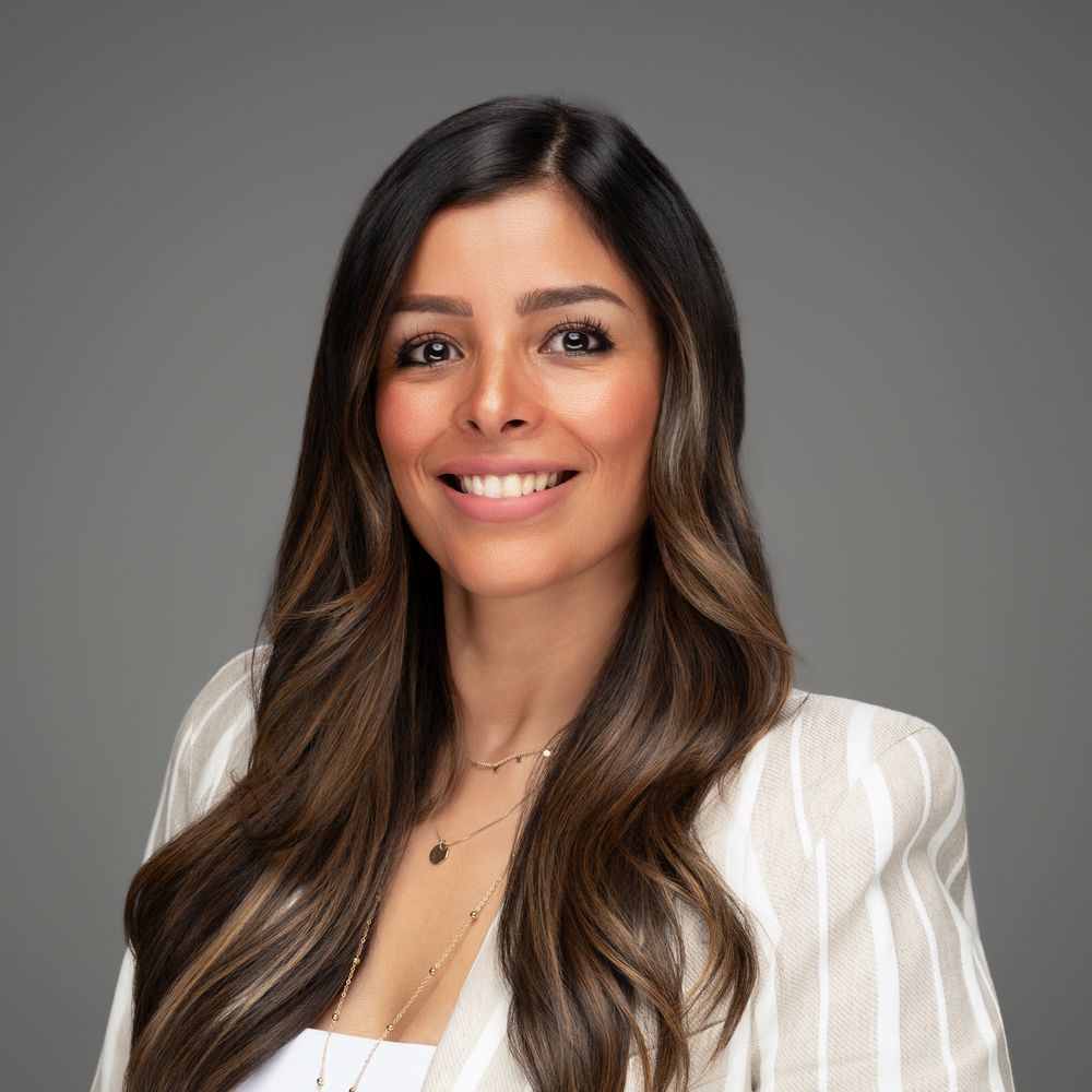 Professional headshot portrait in white blazer against gray background with warm smile and long wavy brown hair.