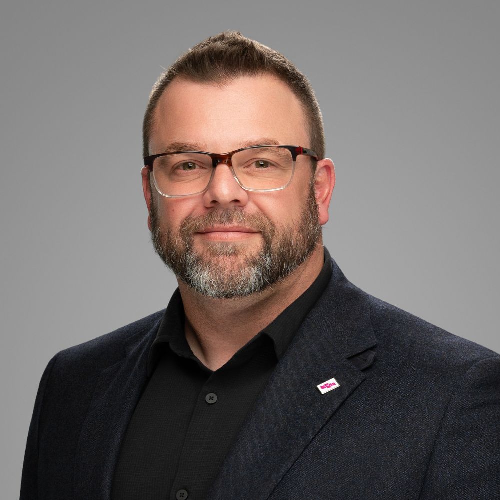 Professional headshot of a person in glasses and dark blazer against a gray background.