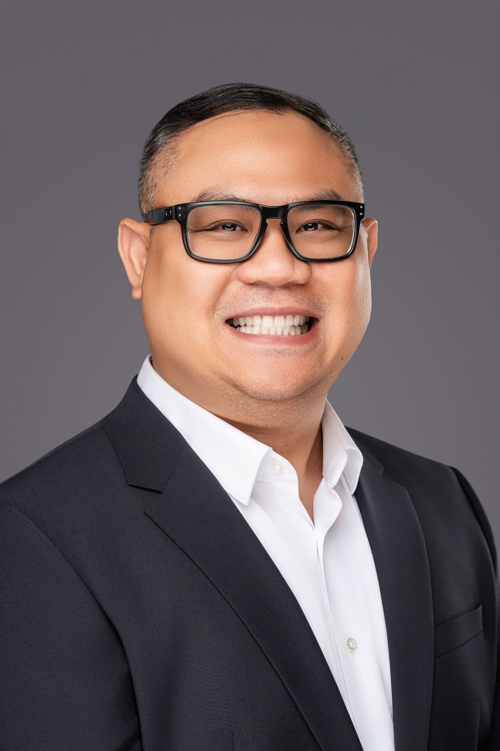 Professional headshot of a person in glasses wearing a black suit jacket and white dress shirt against a gray background.