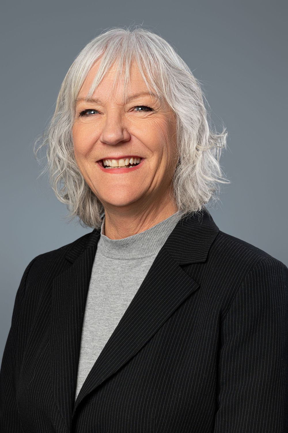 Professional studio portrait of someone in black blazer and gray turtleneck with silver hair against gray backdrop.