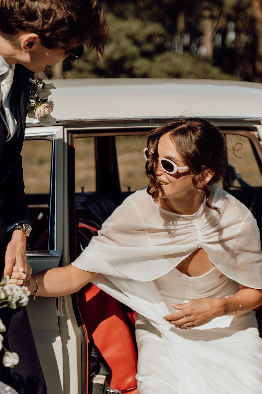 A person in a white dress sits in a vintage car with red interior seats on a sunny day.