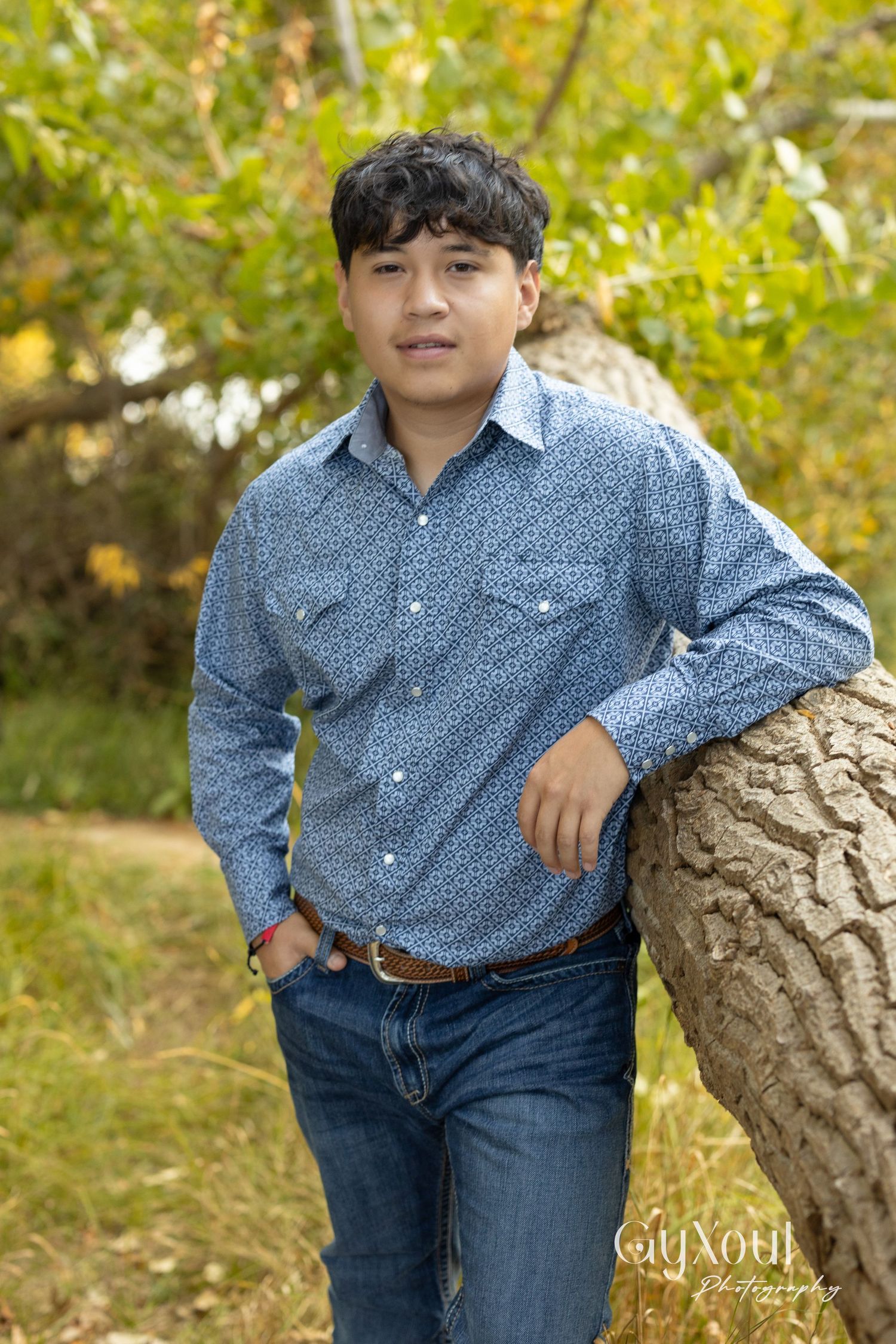 Person in patterned blue button-down shirt and jeans leaning against tree trunk outdoors.