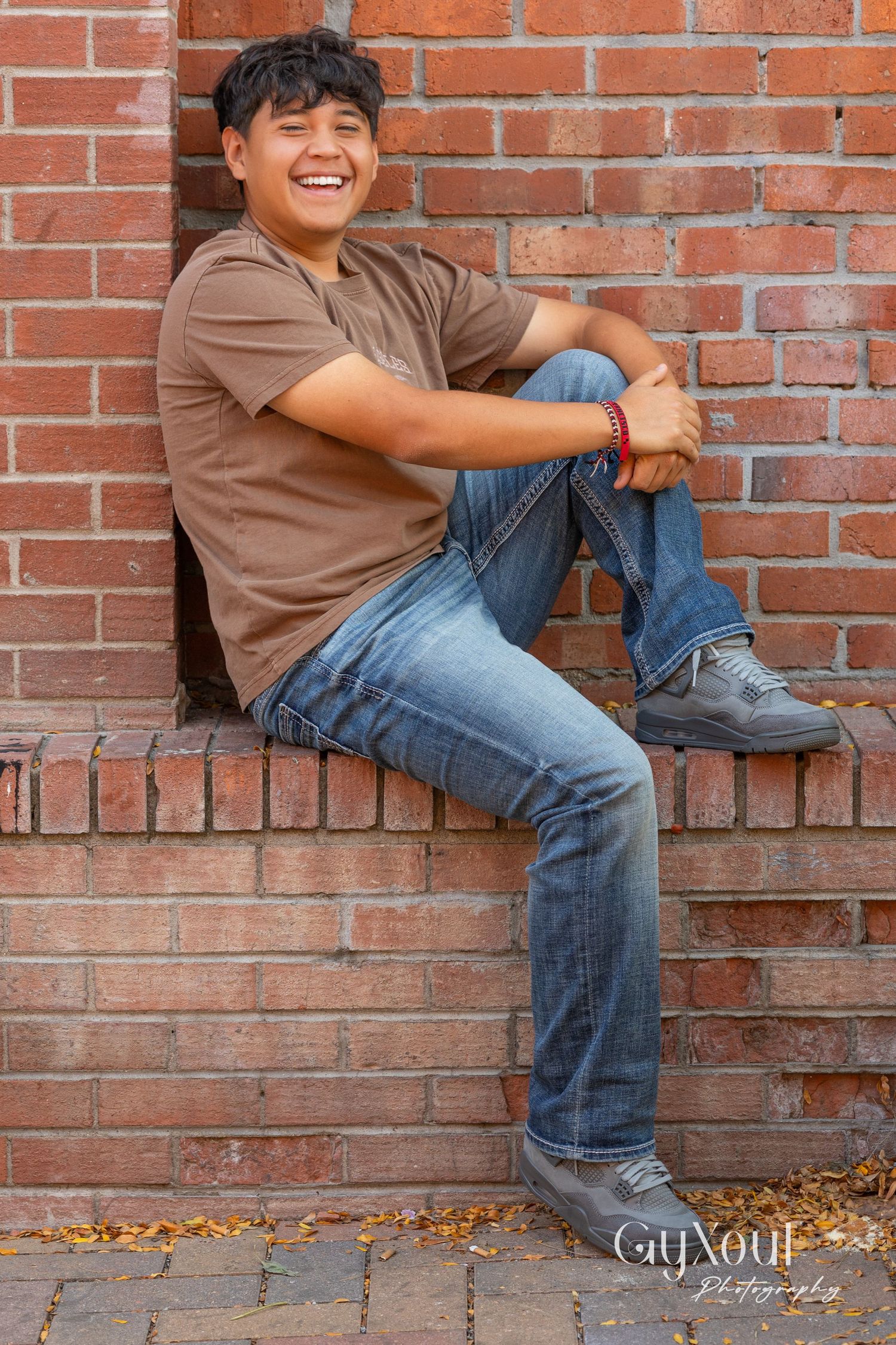 Person in brown t-shirt and jeans sitting on brick ledge against wall outdoors.