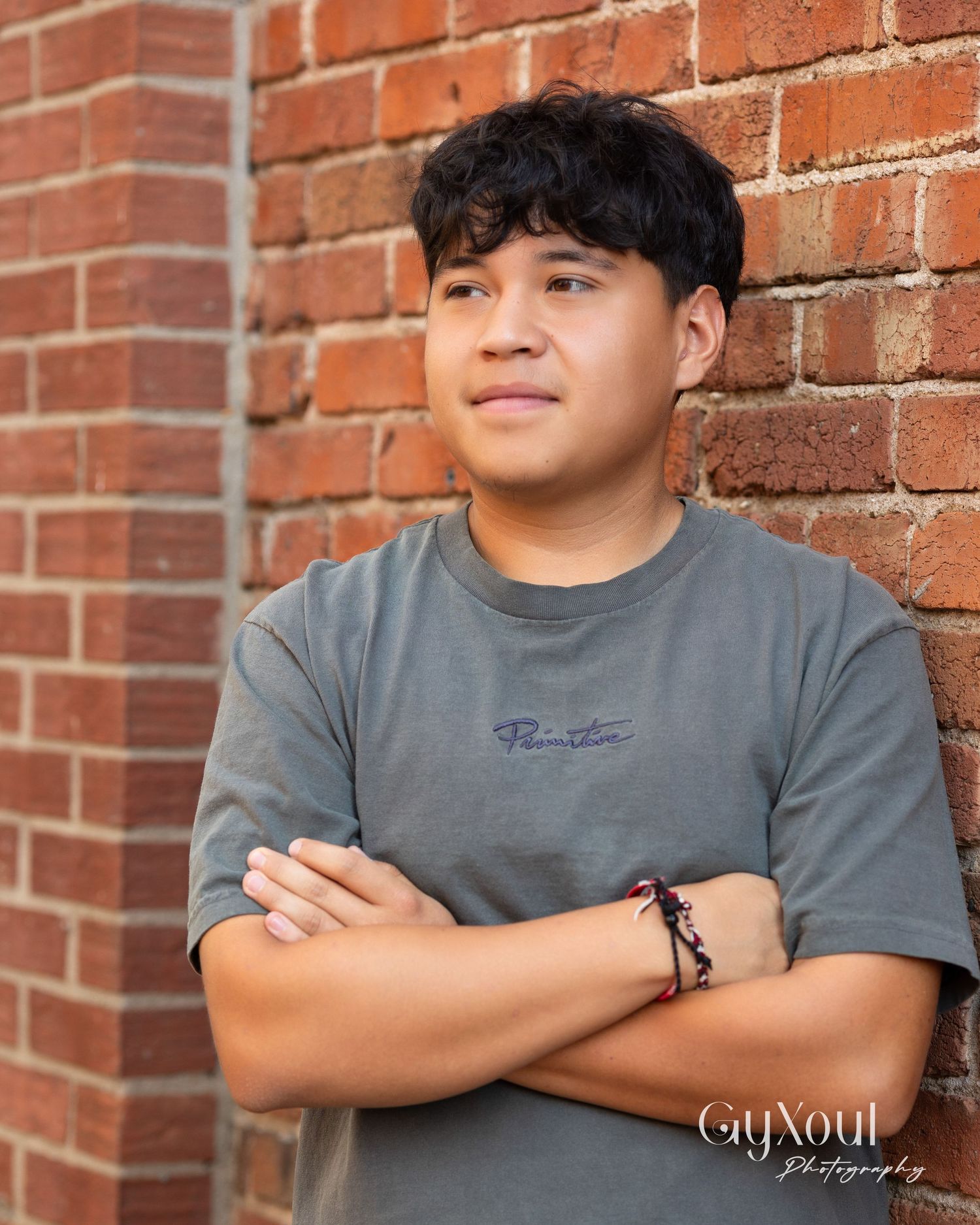 Portrait against red brick wall showing person in grey t-shirt with crossed arms and bracelet.