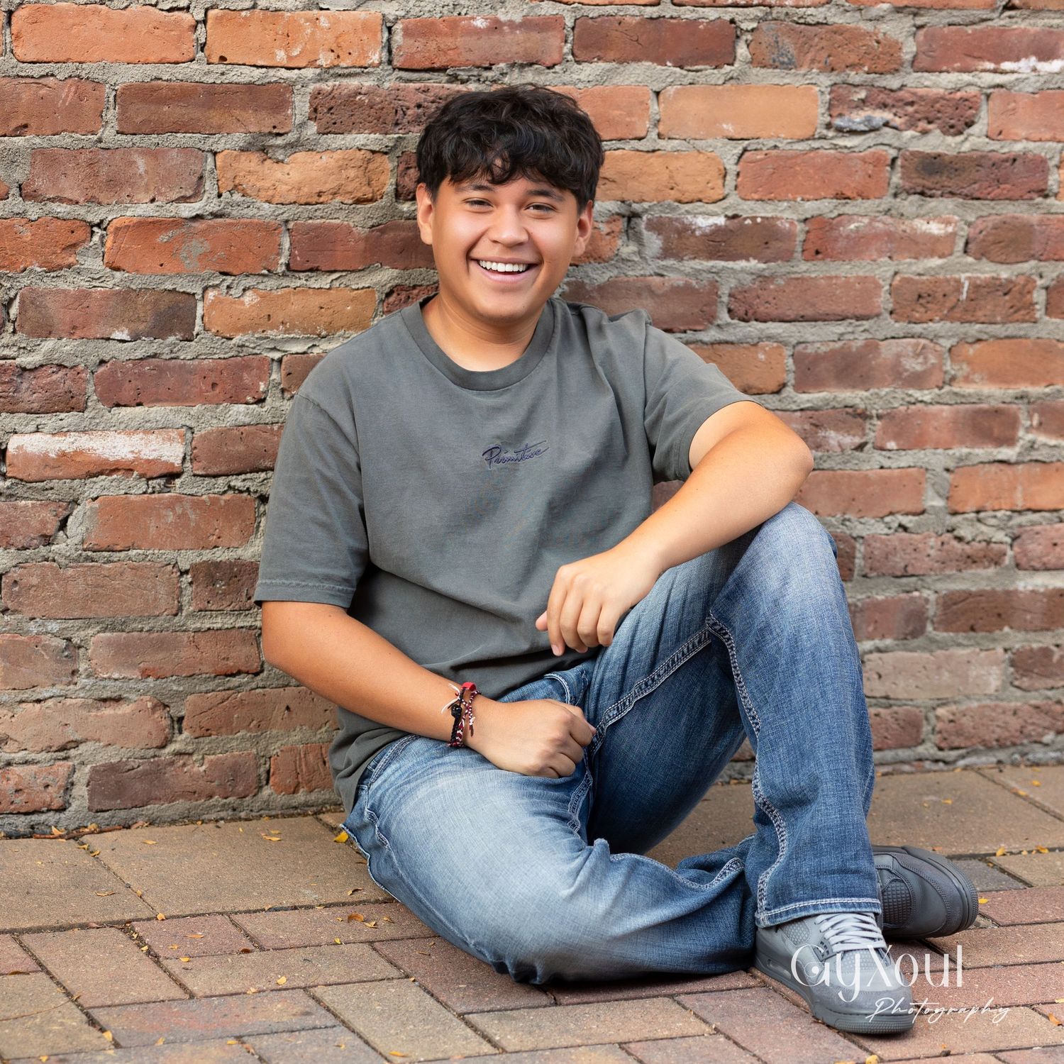 Person in grey t-shirt and jeans sitting casually against brick wall outdoors.