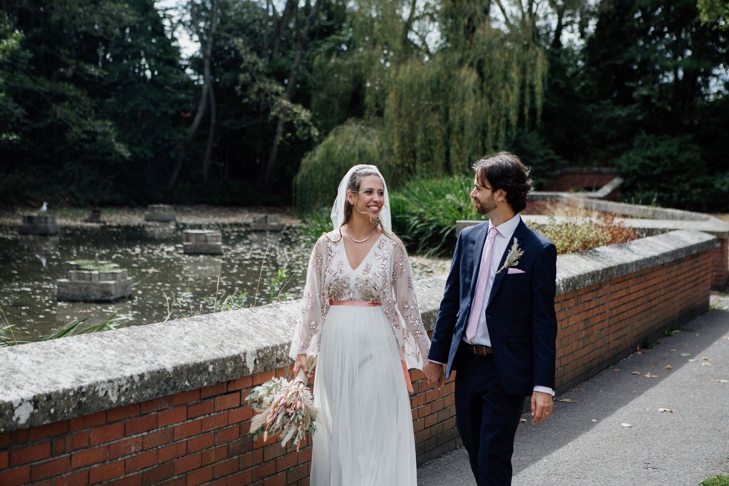 Newlywed couple walks along a brick wall path surrounded by lush greenery on their wedding day.
