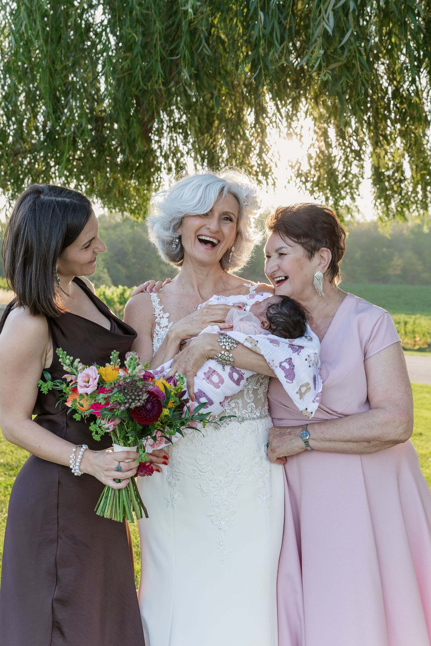 Joyful moment shared between four generations of women under willow trees at a wedding at Vineland Estates Winery.