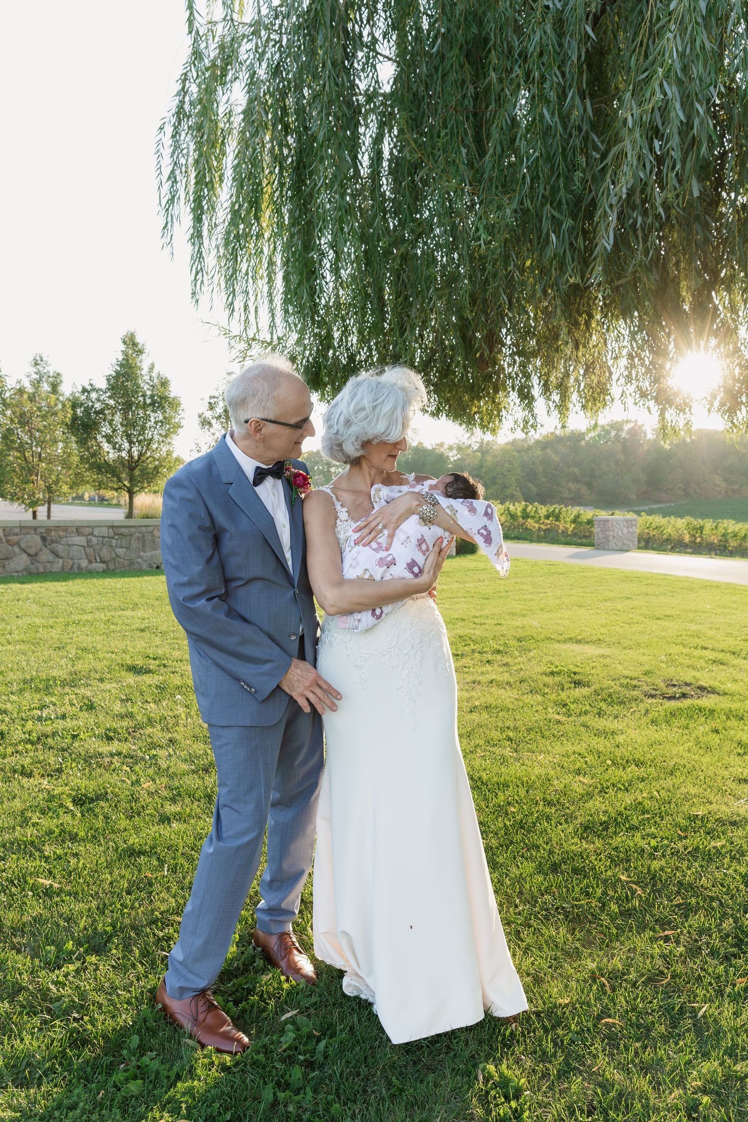 Bride and groom hold their newborn granddaughter at sunset under a willow tree at Vineland Estates Winery.