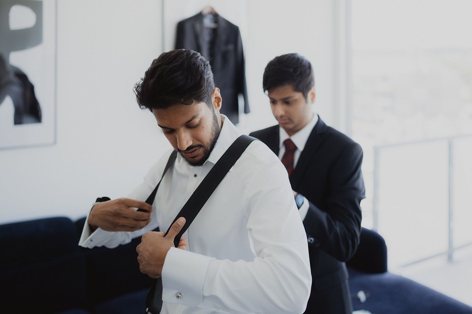Groom in black tuxedo adjusts his bow tie while getting ready in a minimalist white room.