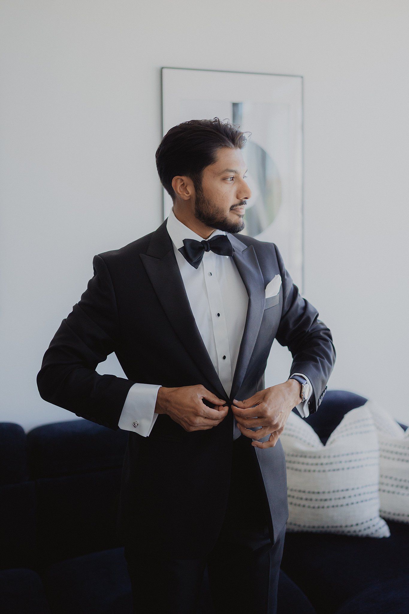 Person in formal black tuxedo adjusts cufflinks while seated in elegant room.
