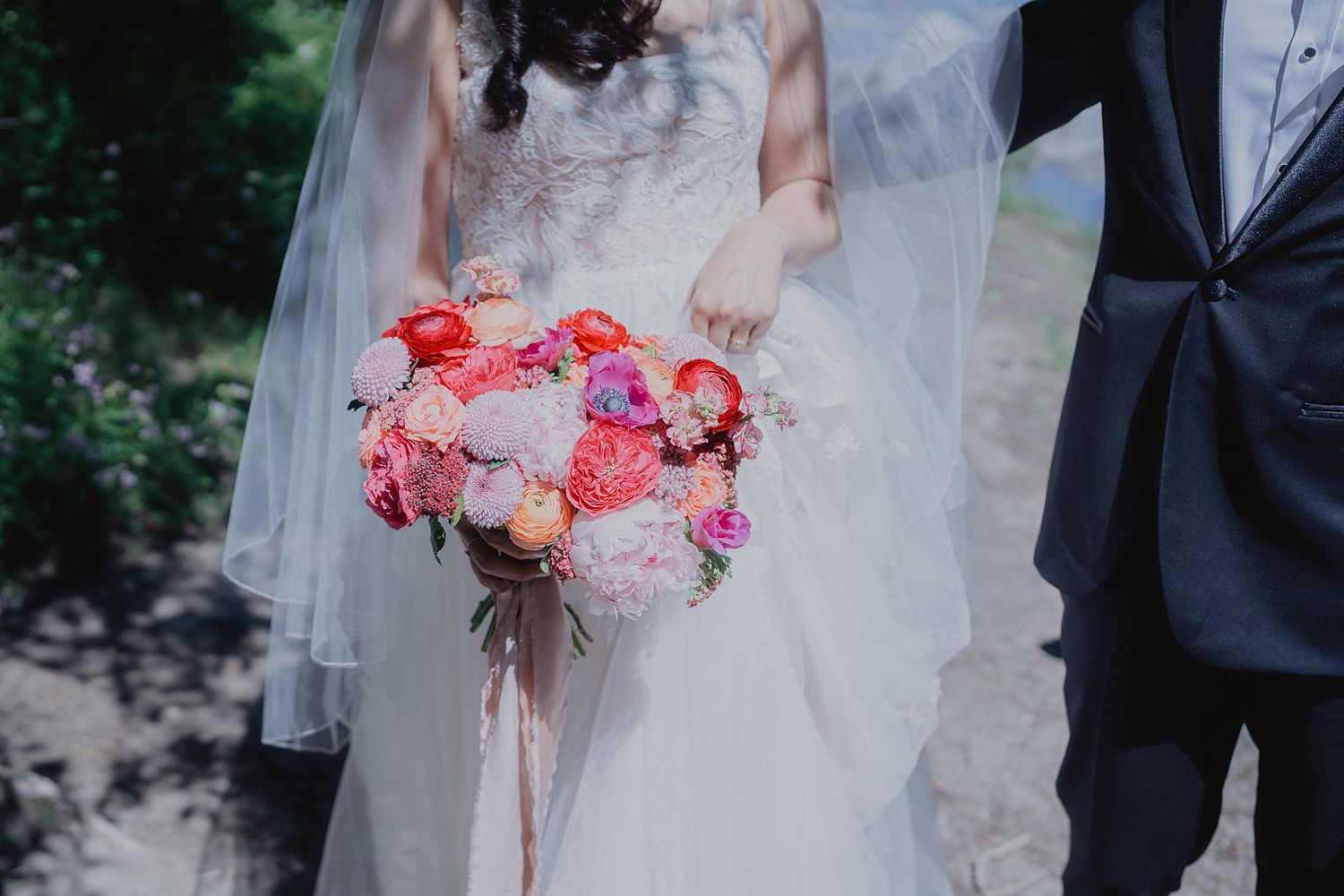 A romantic wedding couple poses in a lush garden setting holding vibrant coral and pink bouquets.