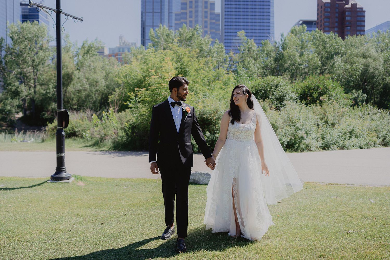 Wedding couple strolling along a paved path with city skyline and green trees in the background.
