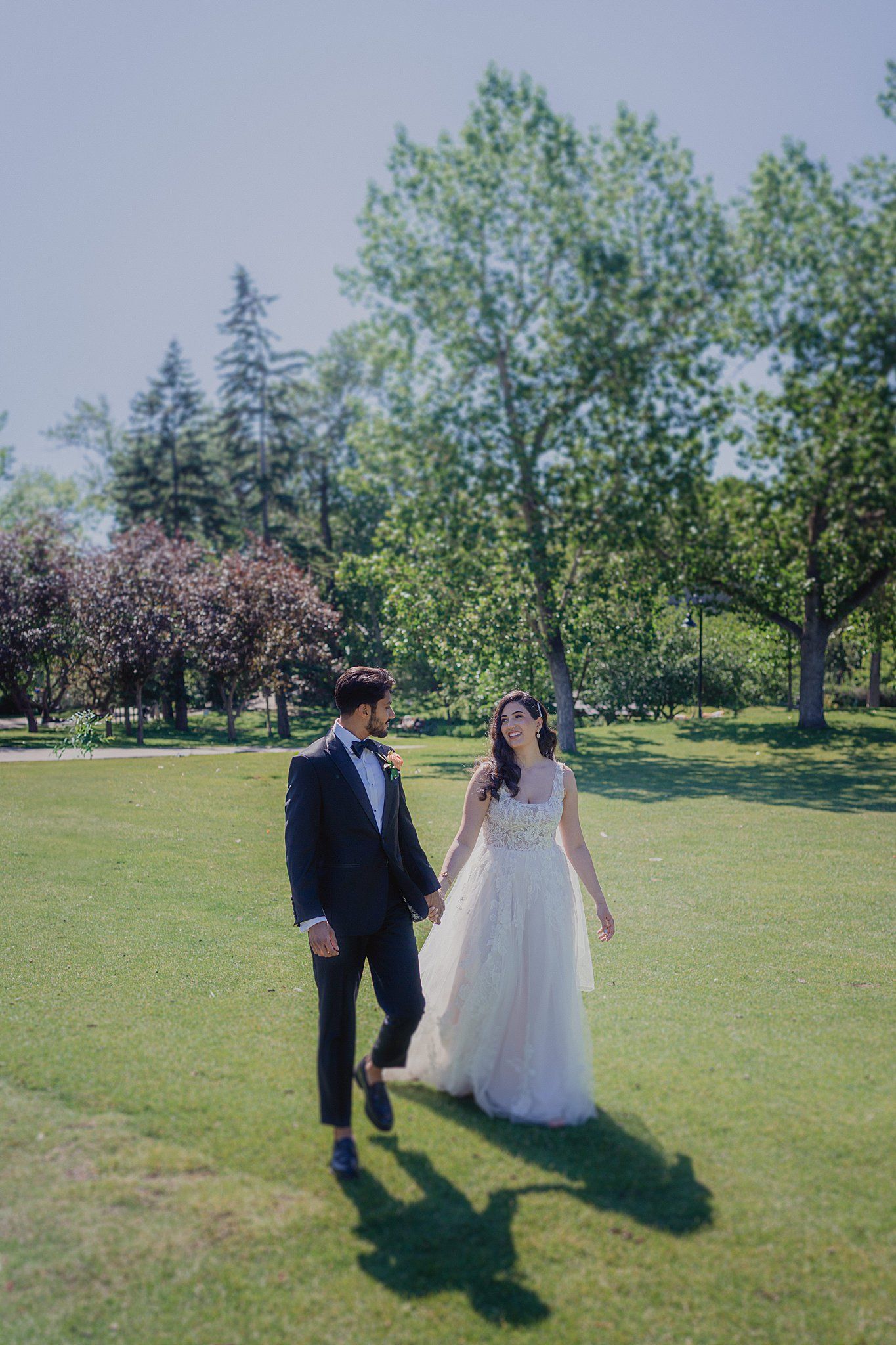 A couple walks together on a sunny grassy lawn surrounded by blooming trees in a romantic outdoor setting.