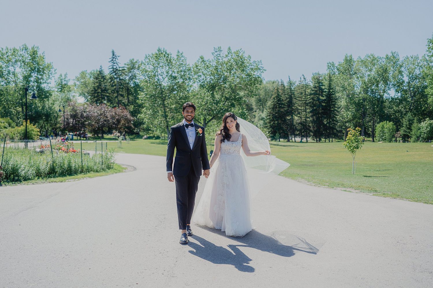 A sunny wedding photoshoot on a paved pathway surrounded by green summer parkland.