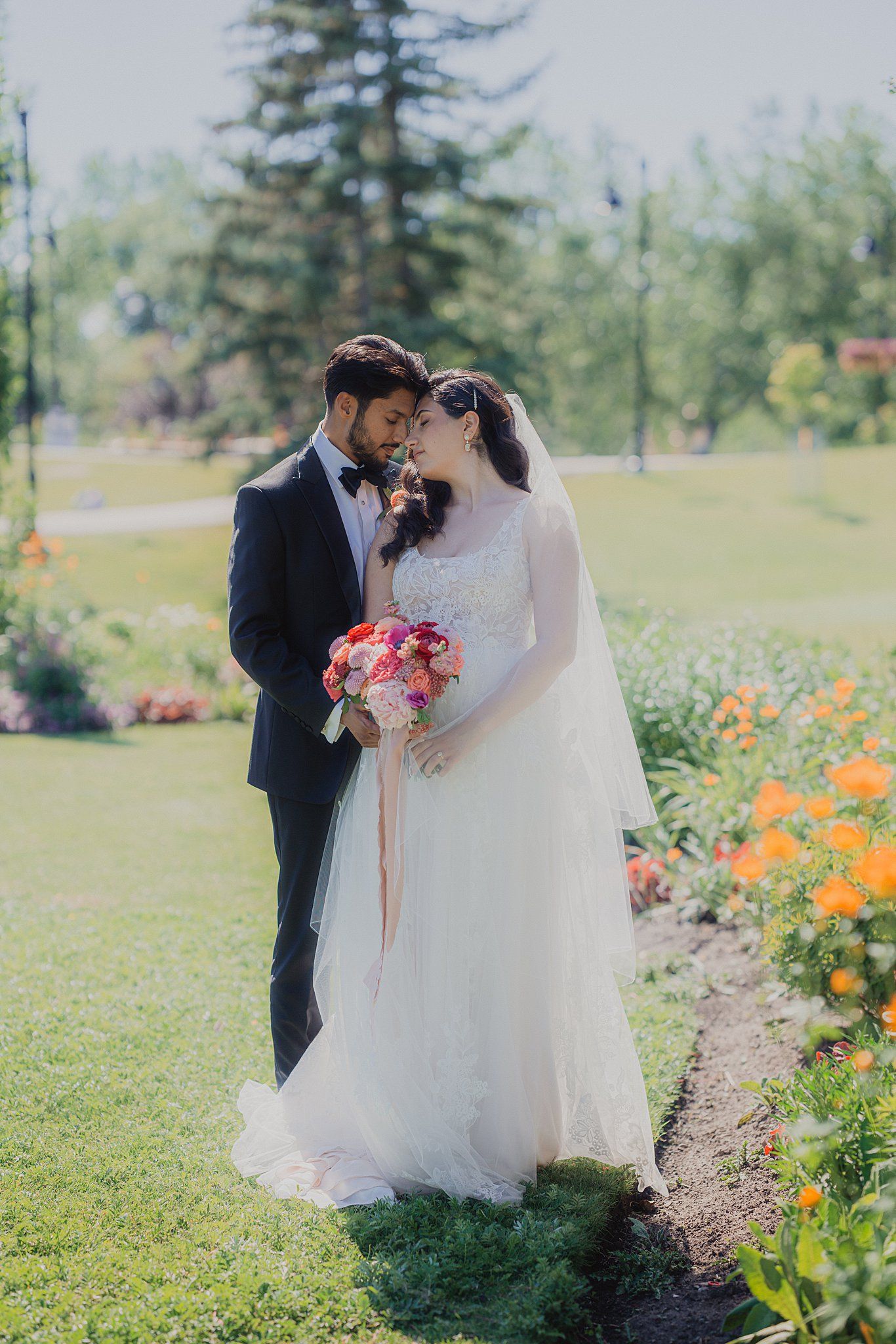 Romantic wedding couple poses in a garden filled with colorful flowers and evergreen trees on a sunny day.
