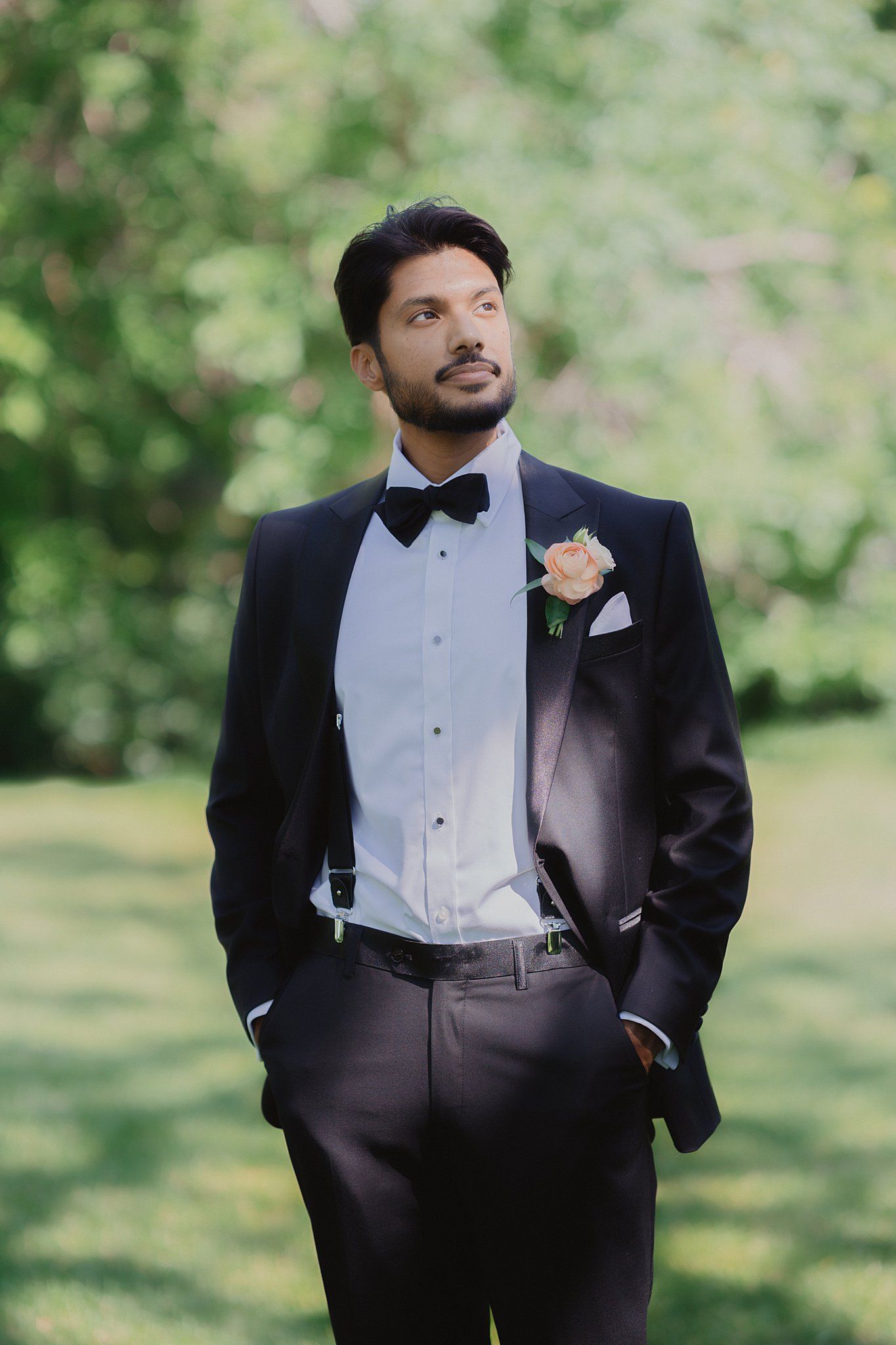 Groom in classic black tuxedo and white dress shirt poses in a garden setting.