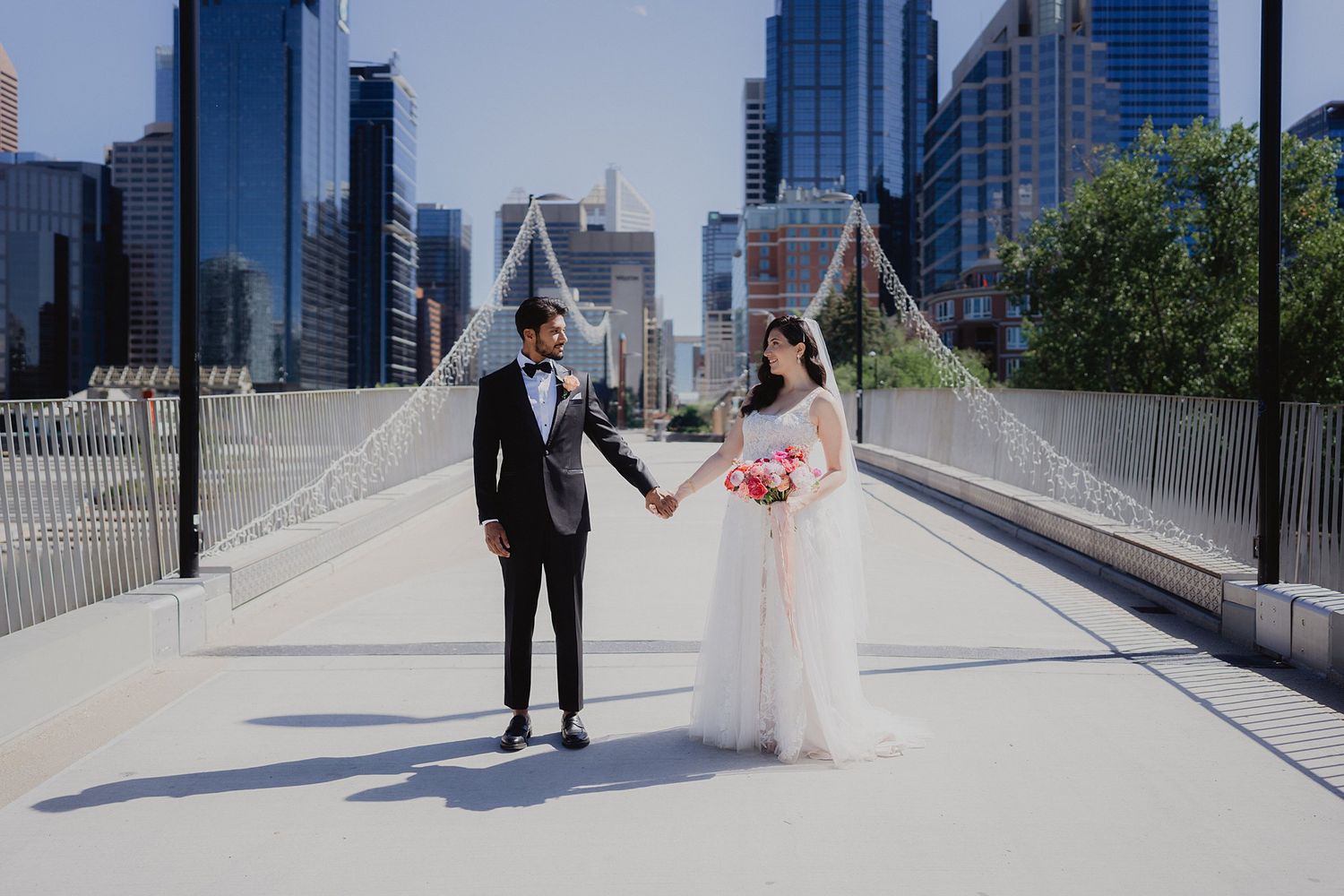 Newlyweds pose against modern city skyline backdrop on sunny day.