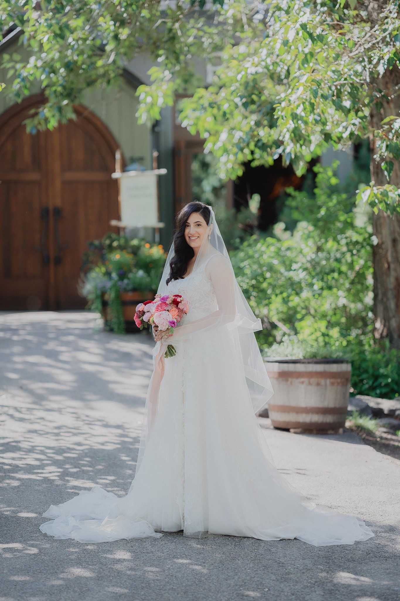 Bride in flowing white wedding gown poses elegantly on a stone pathway near wooden barrels.