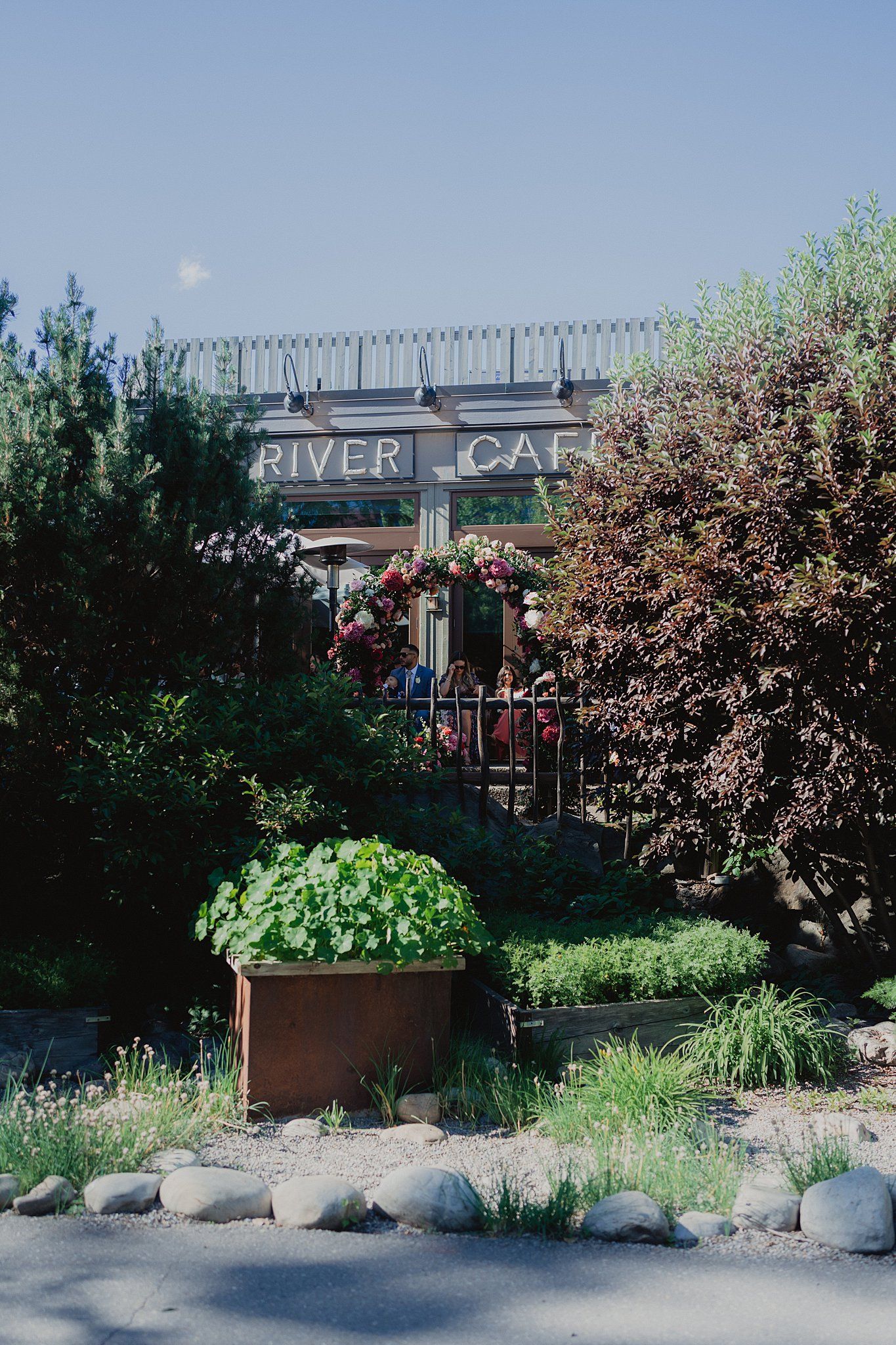 River Cafe exterior with landscaped garden beds, decorative rocks and flowering trees along the entrance.