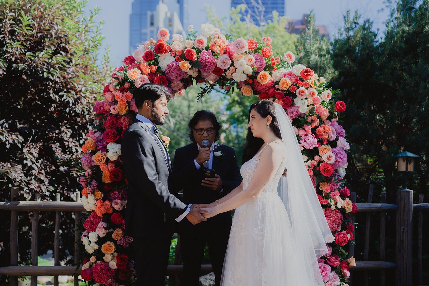 Wedding ceremony setup with vibrant pink and red floral arch arrangements in an outdoor garden setting.