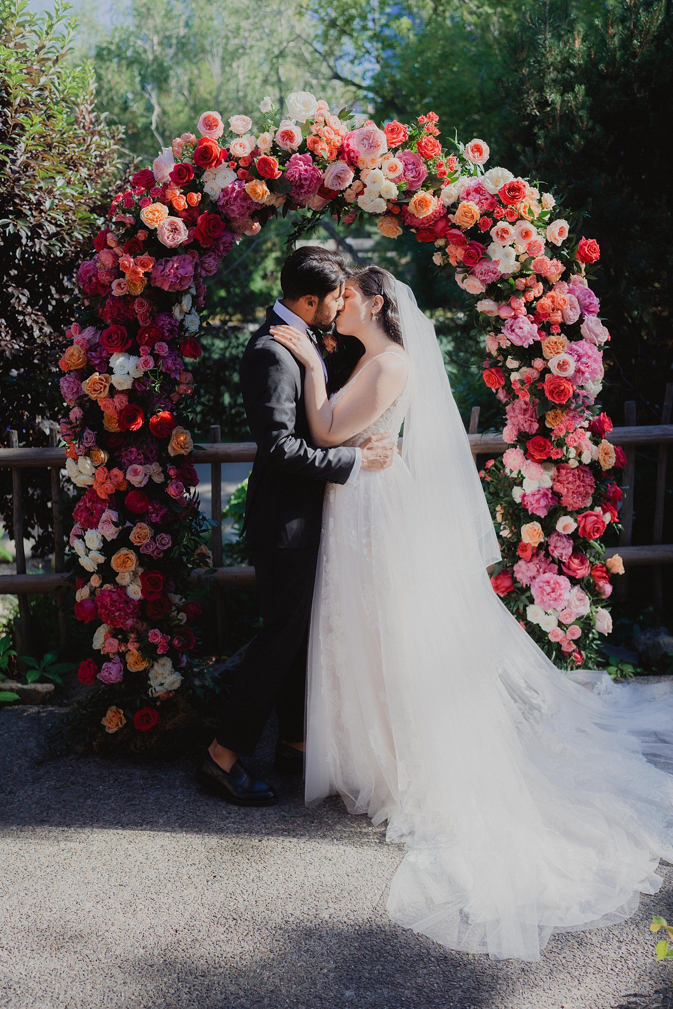 Romantic wedding couple embraces under a stunning floral arch with pink and red roses in an outdoor garden setting.