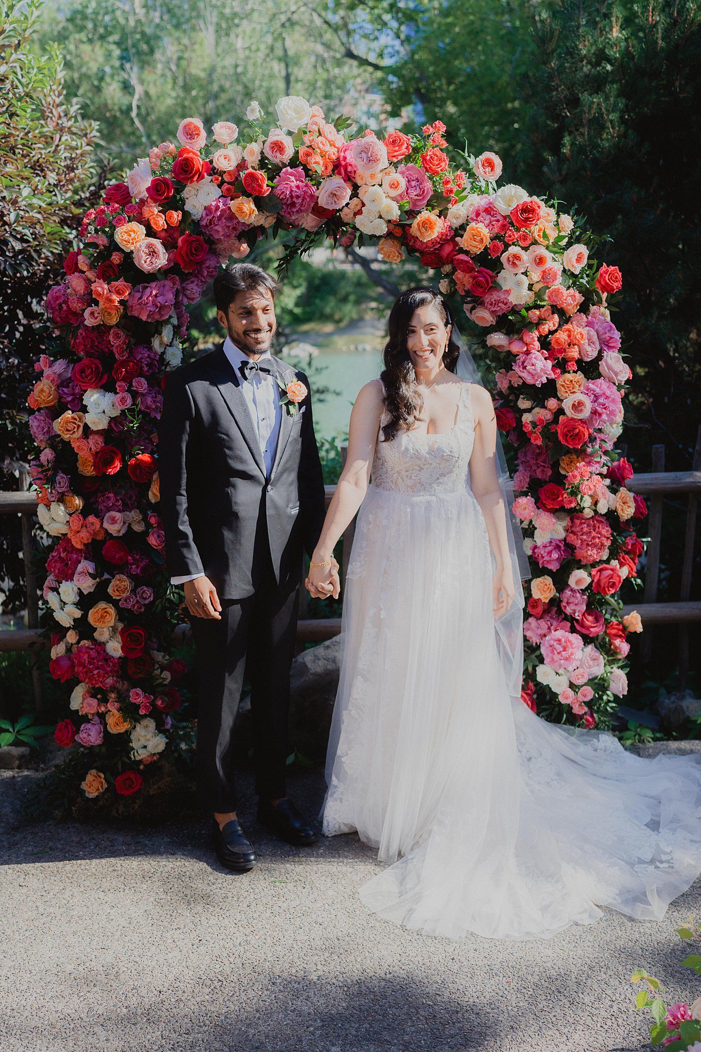Wedding ceremony setup with a stunning floral arch featuring pink and coral roses in a garden setting.