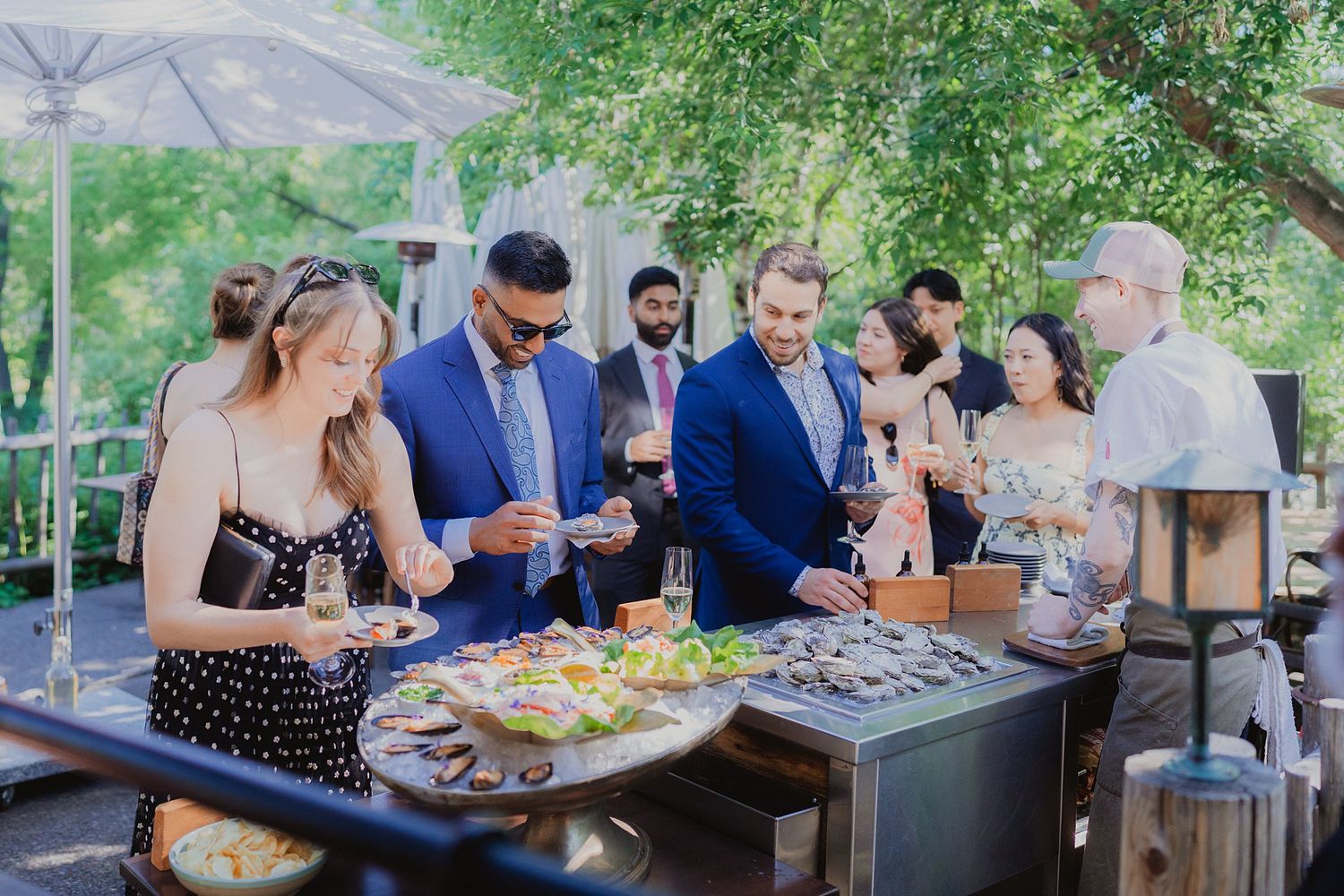 Wedding guests gather around an outdoor buffet table under a canopy on a sunny day.
