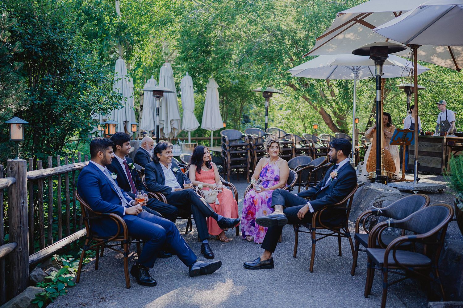 Wedding guests enjoying an outdoor reception on a patio with umbrellas and tables in a garden setting.