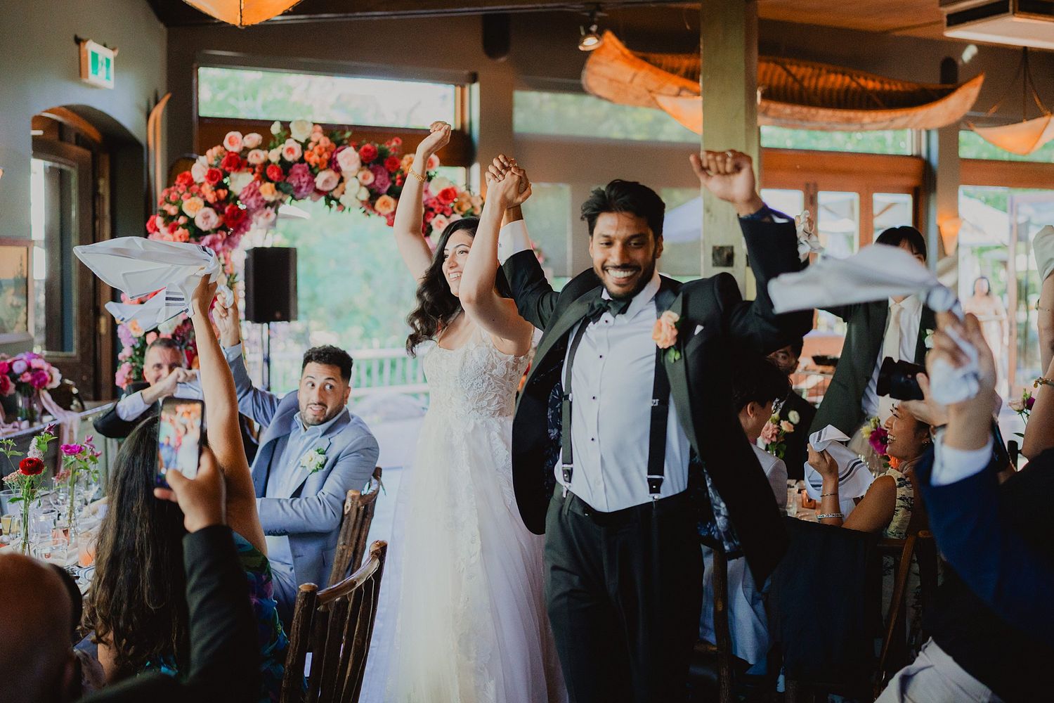 Wedding celebration in a bright indoor venue with guests dancing and mingling around decorated tables.
