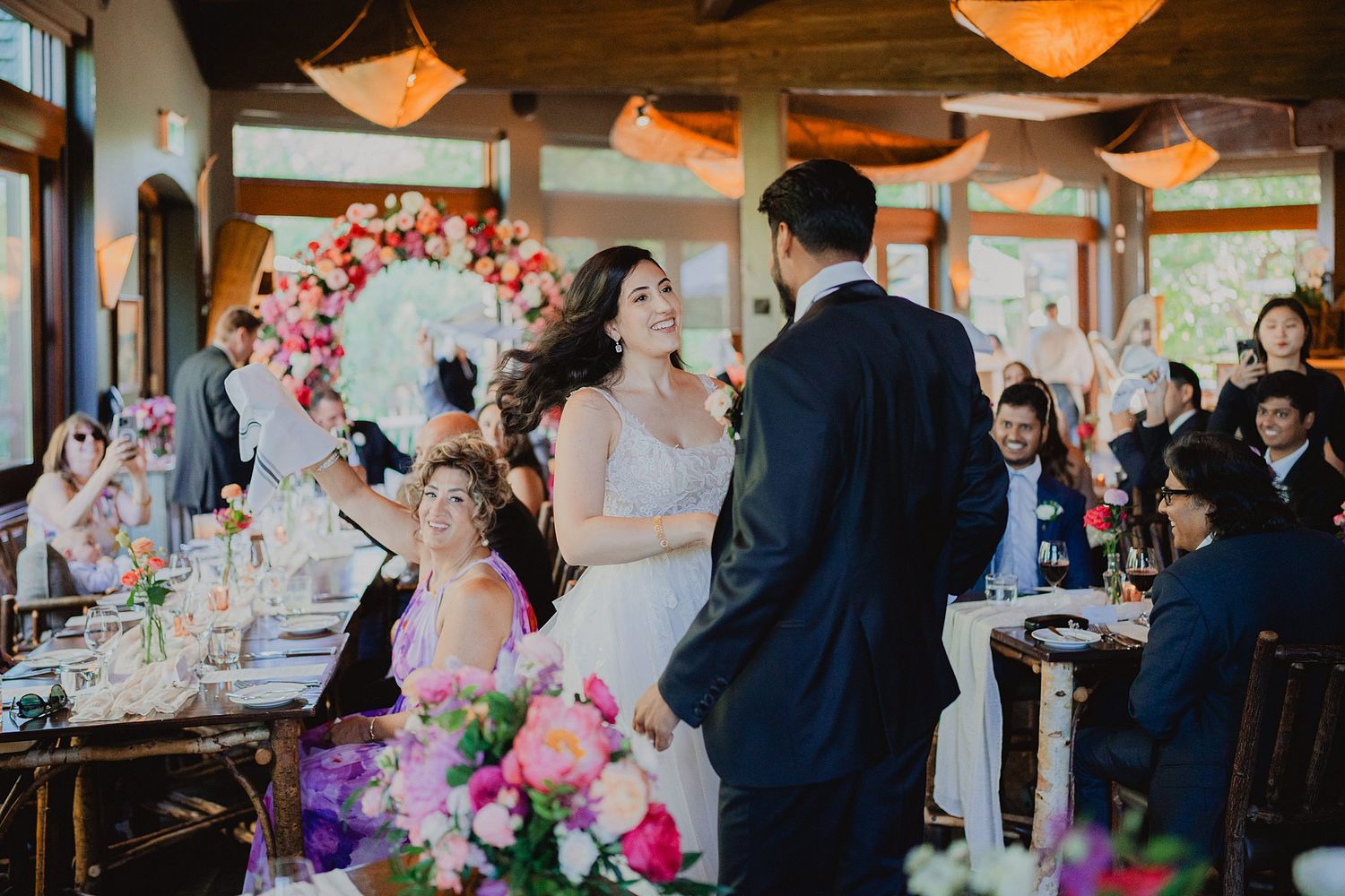 Wedding reception guests enjoying dinner at elegantly decorated tables with hanging pendant lights and floral centerpieces.