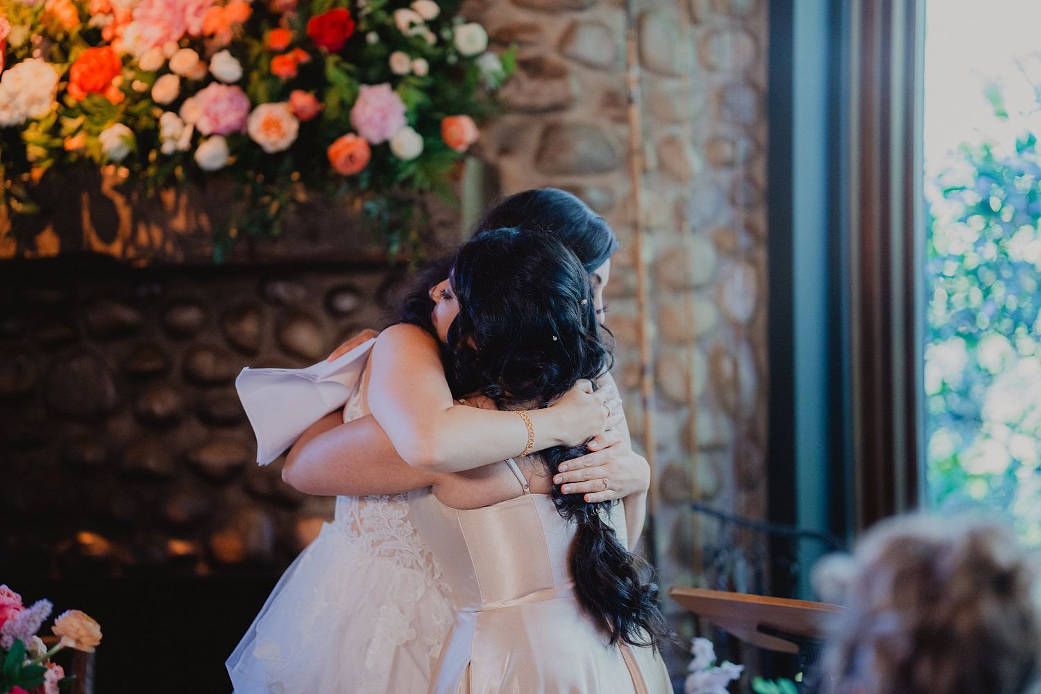 Two people share an emotional embrace during a wedding celebration with floral decorations in the background.