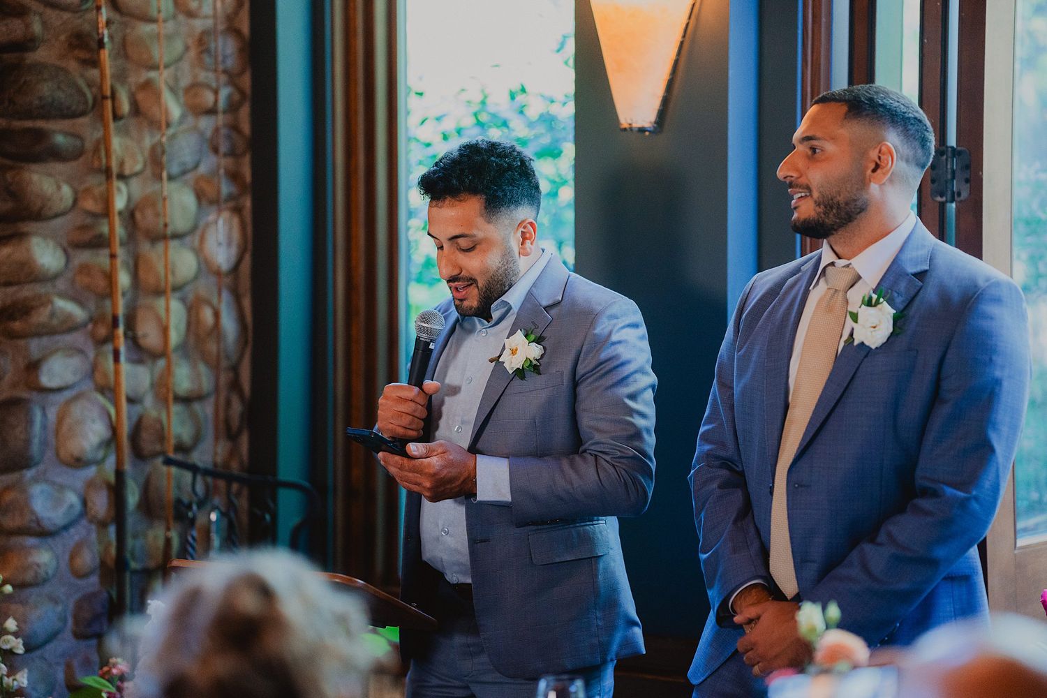 Two groomsmen in formal attire share a lighthearted moment during wedding speeches.