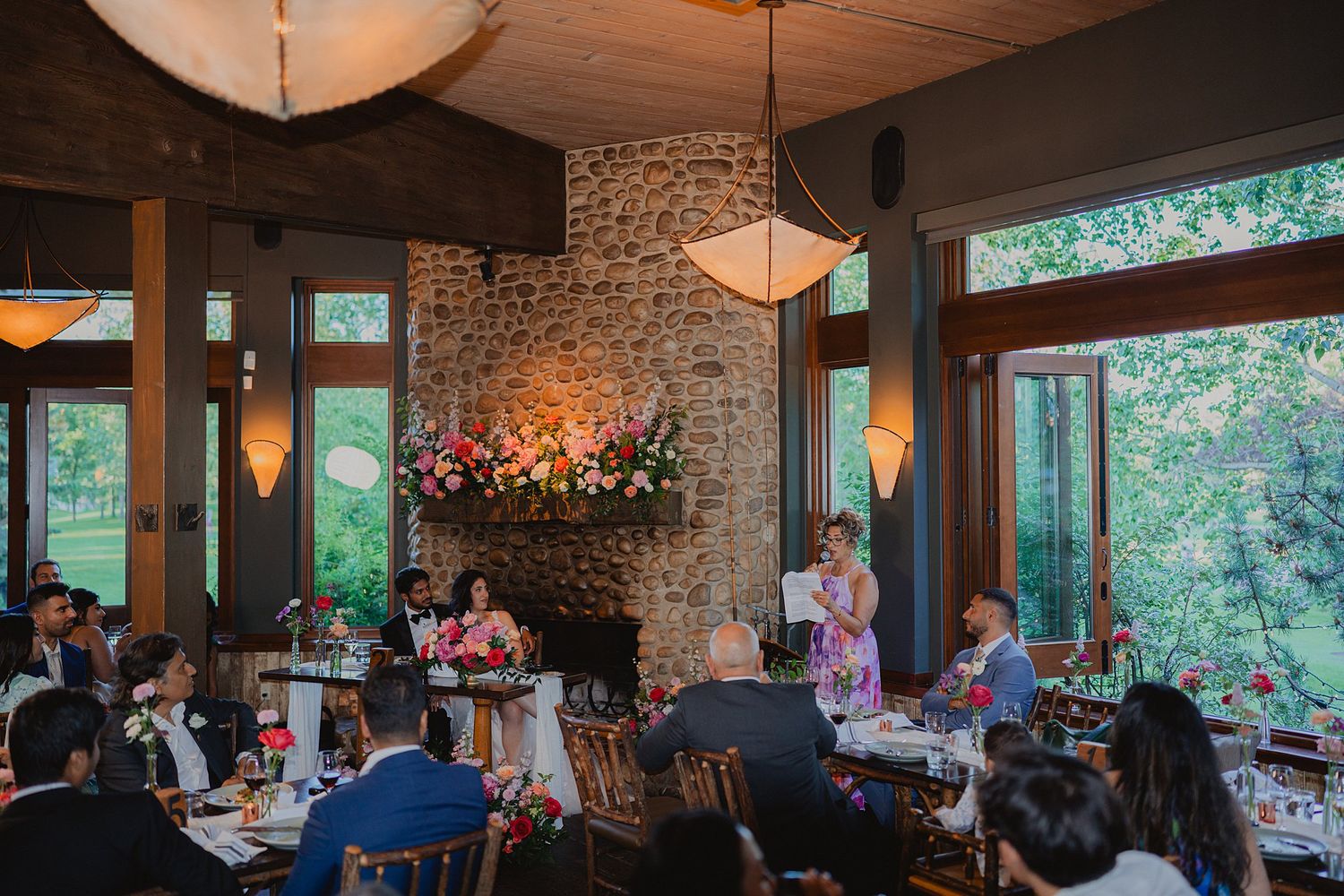 Wedding reception in a rustic stone-walled venue with pendant lighting and forest views.