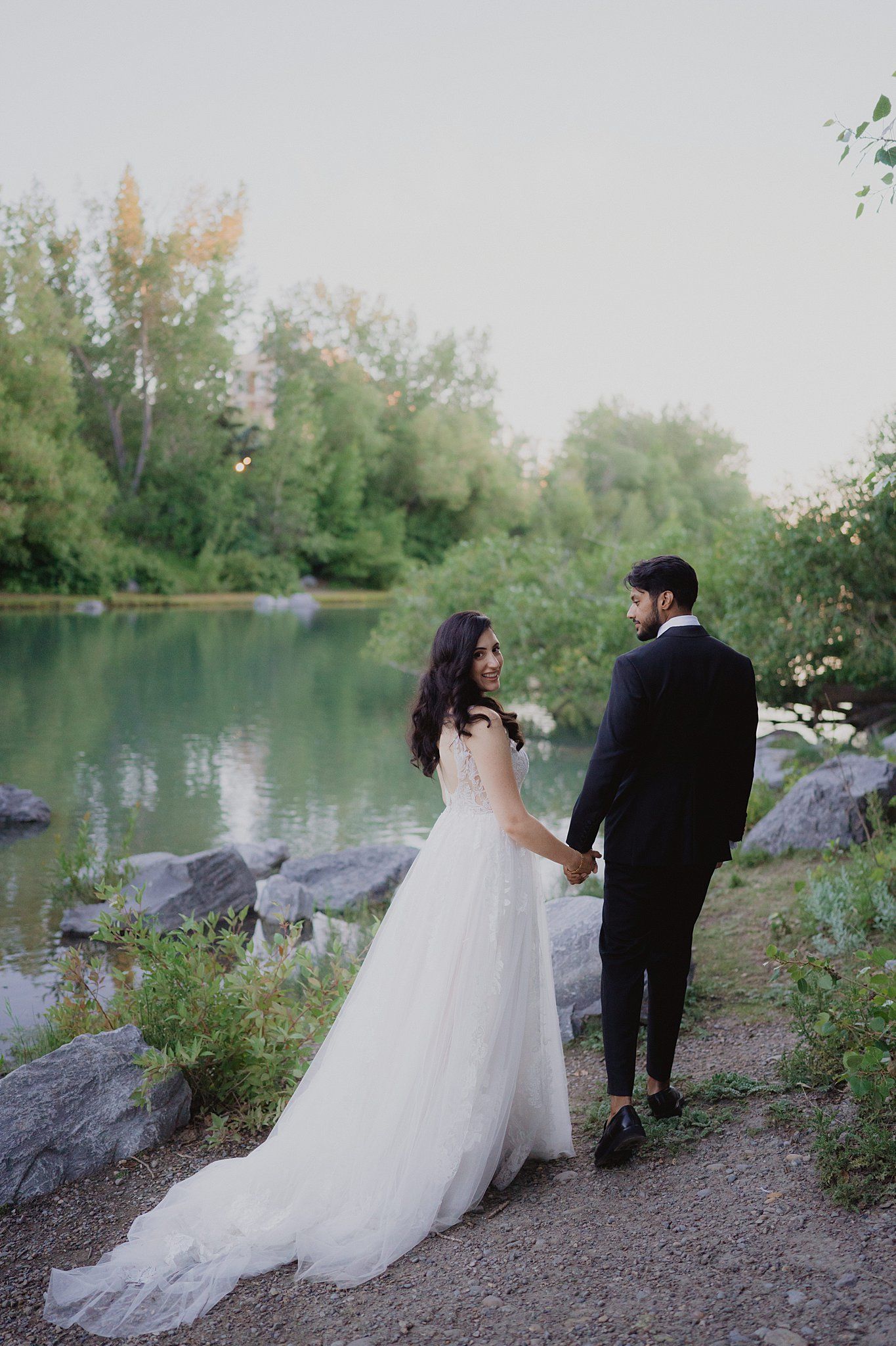 Couple takes romantic photos by a serene lake with white dress trailing on rocky shore.