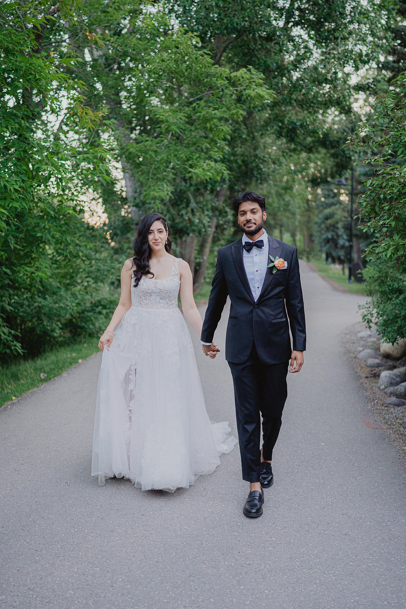 A couple in wedding attire walks along a tree-lined path during a romantic outdoor photo session.