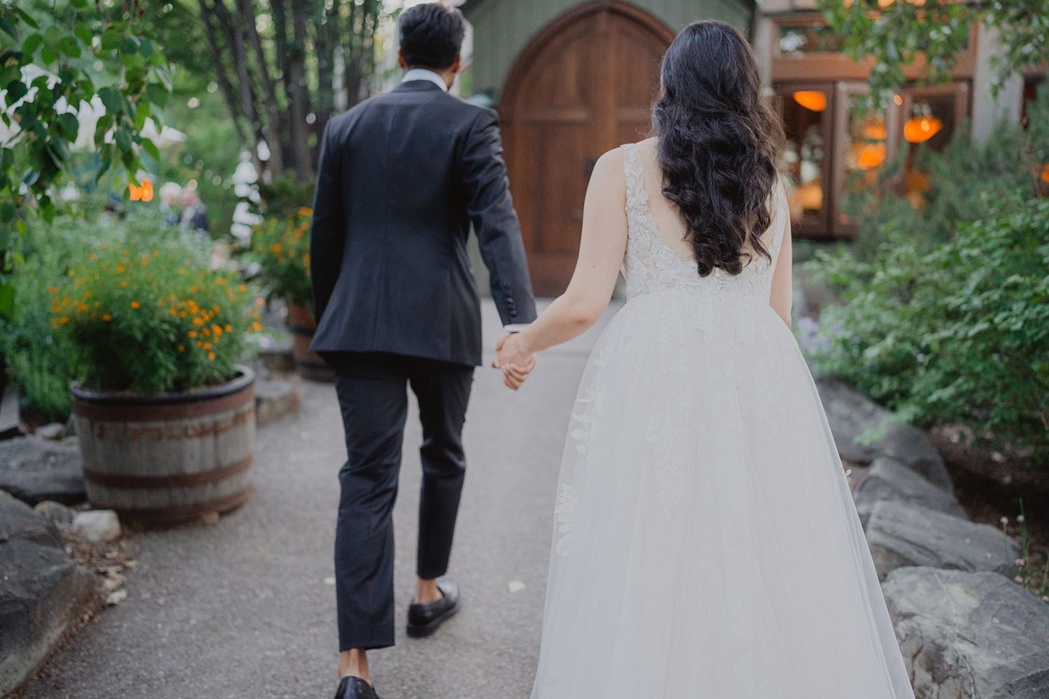 A couple in wedding attire walks hand in hand down a garden path surrounded by greenery.