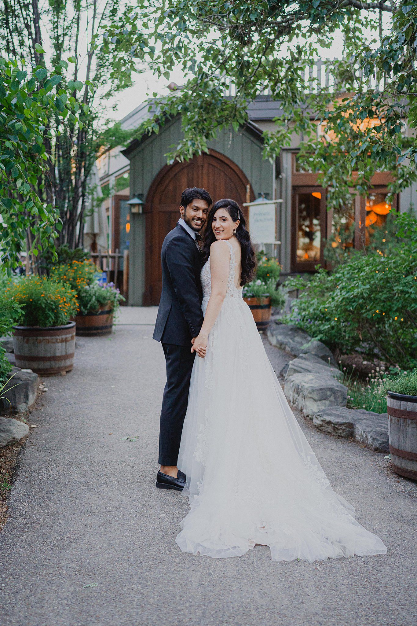A romantic wedding photo of a couple posing on a garden path surrounded by lush greenery and rustic wooden doors.