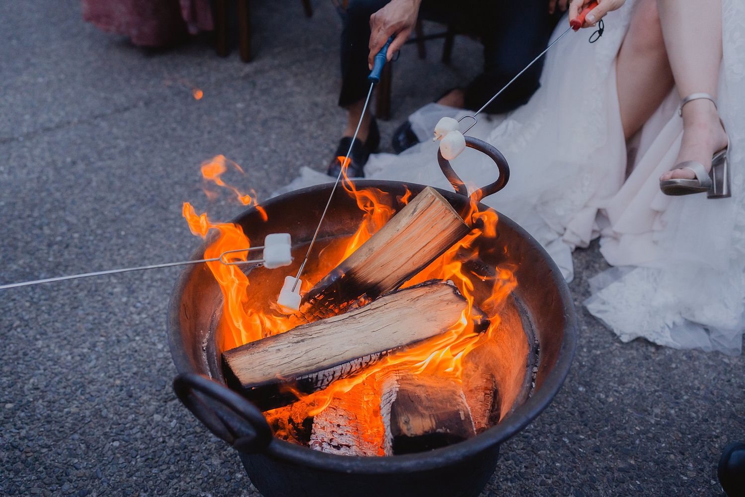 Fire pit with roasting marshmallows glows orange against dark pavement at outdoor wedding reception.