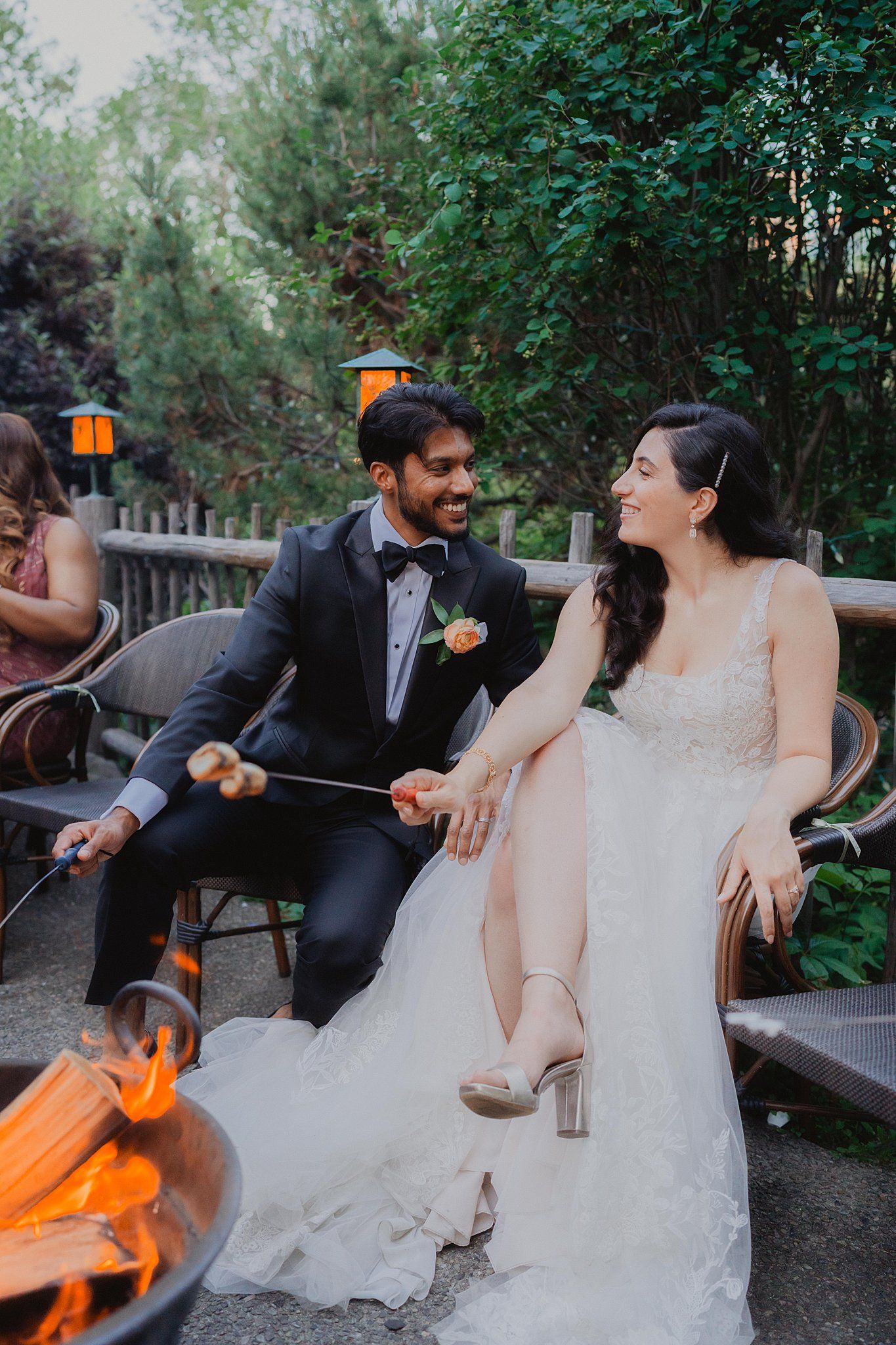 Couple sits on outdoor bench roasting marshmallows during wedding celebration.