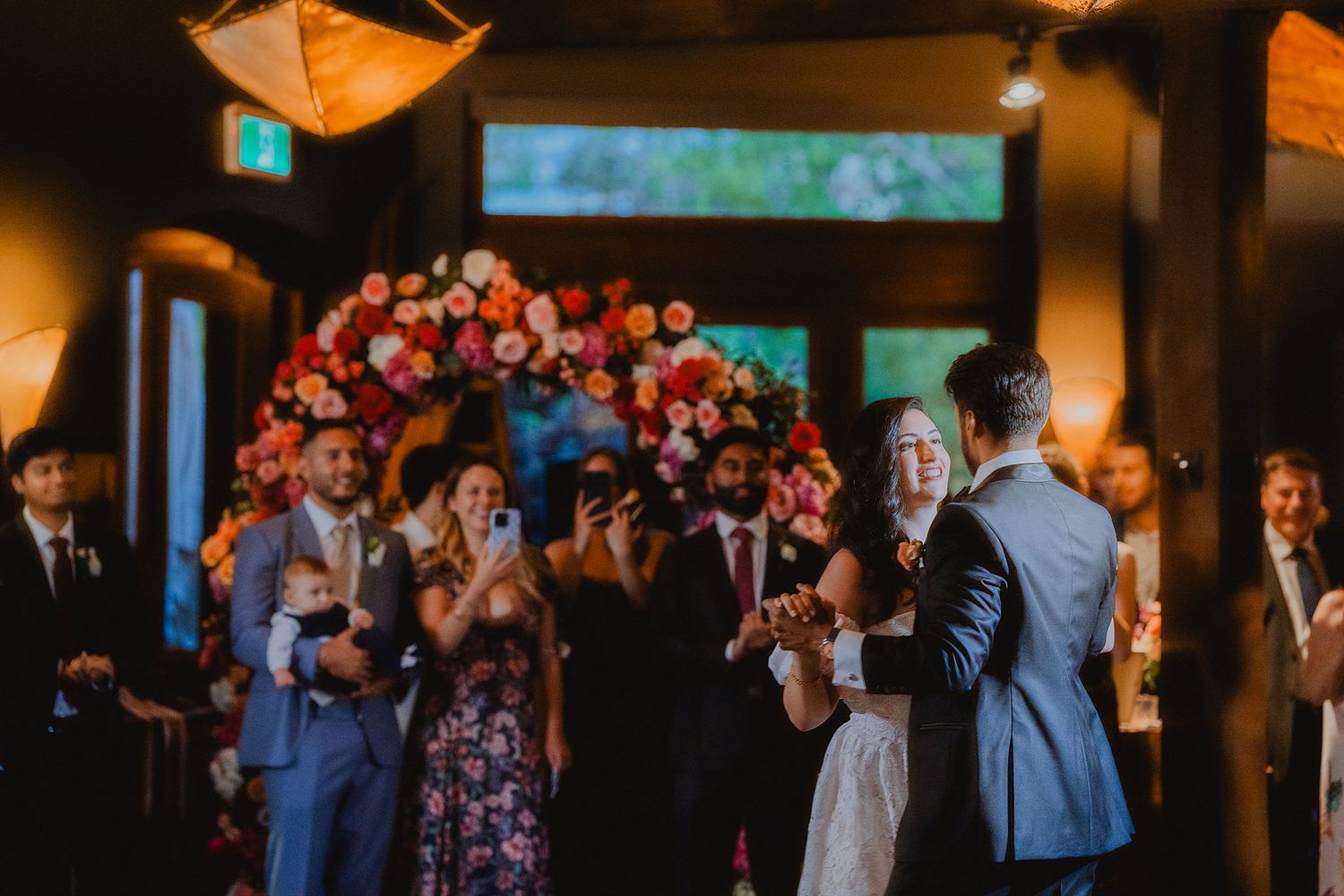 Wedding guests celebrate and cheer during an indoor reception with warm lighting and floral decorations.