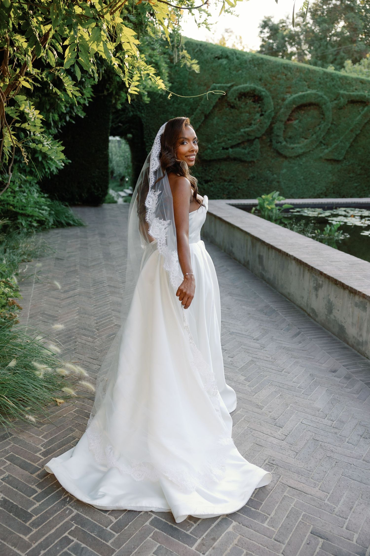 A bride in a flowing white wedding gown walks along a stone pathway surrounded by lush greenery and hedges.