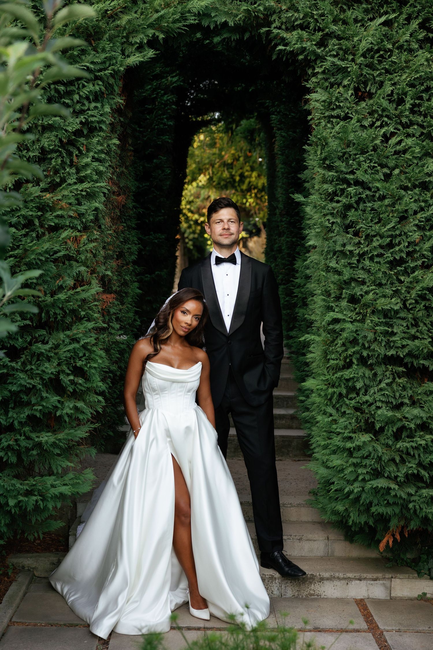 A romantic wedding photoshoot in a garden path lined with lush green hedges.