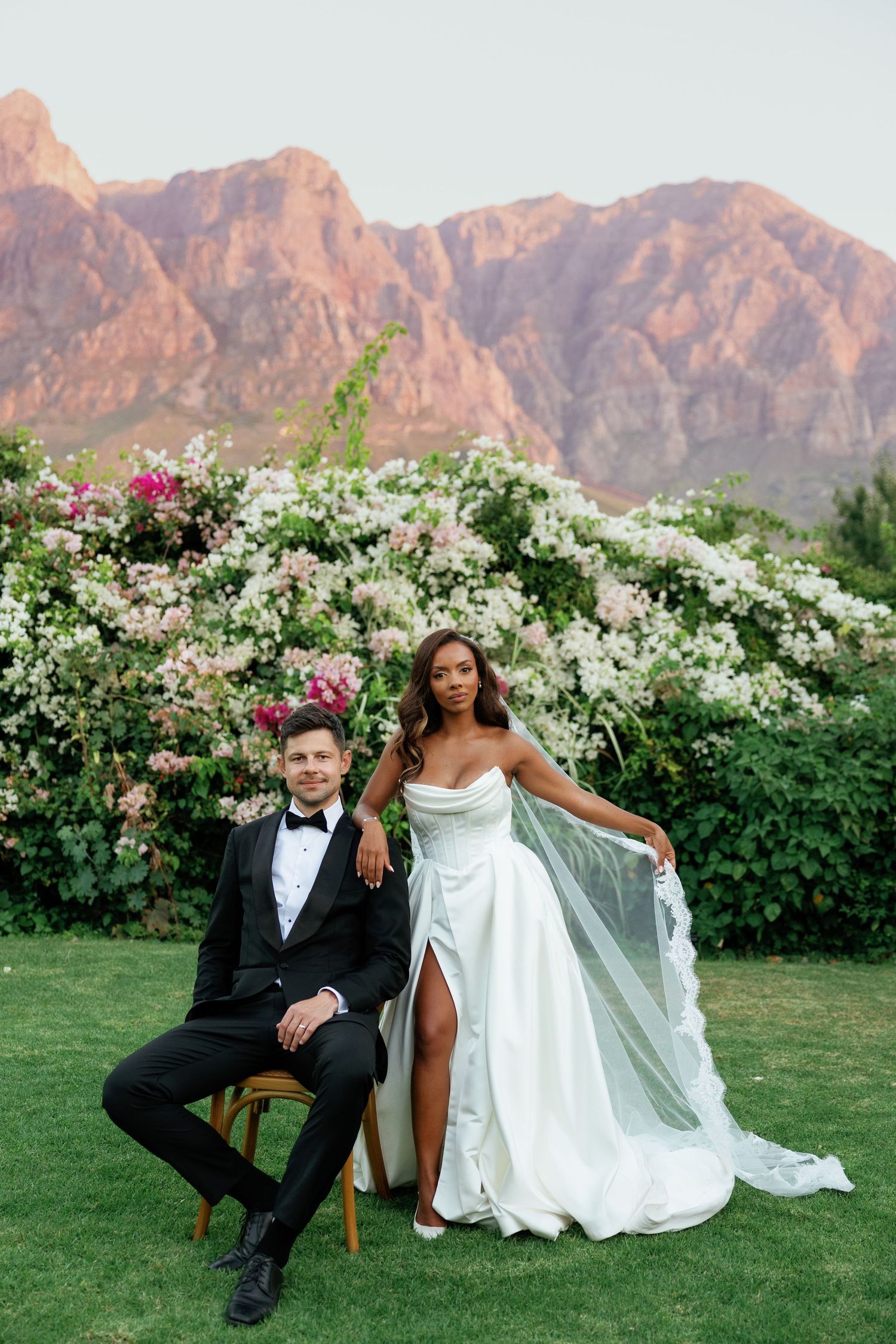 An elegant wedding photo session on a lawn with dramatic mountain backdrop and flowering bushes.