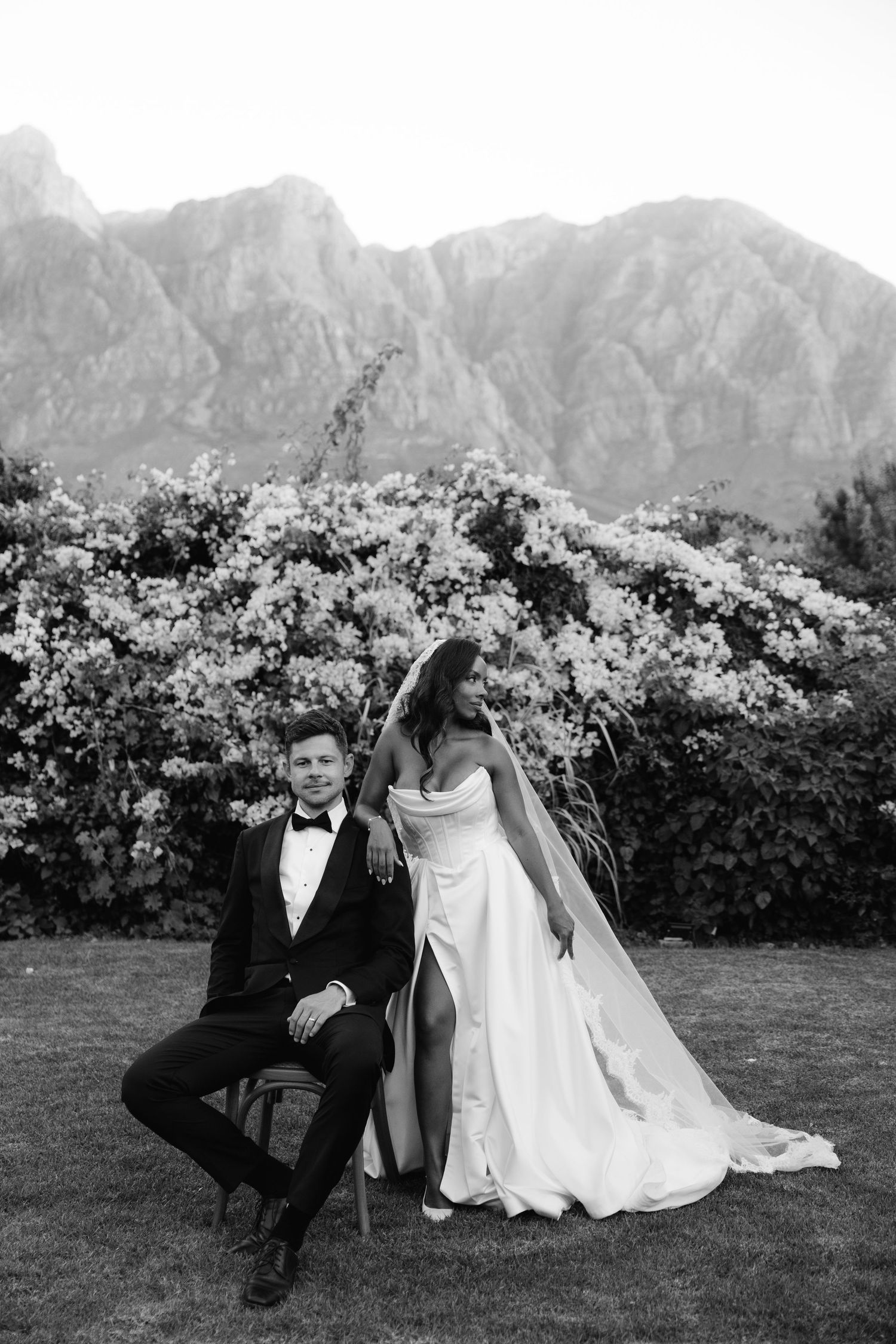 A black and white wedding portrait with majestic mountains and blooming shrubs in the background.