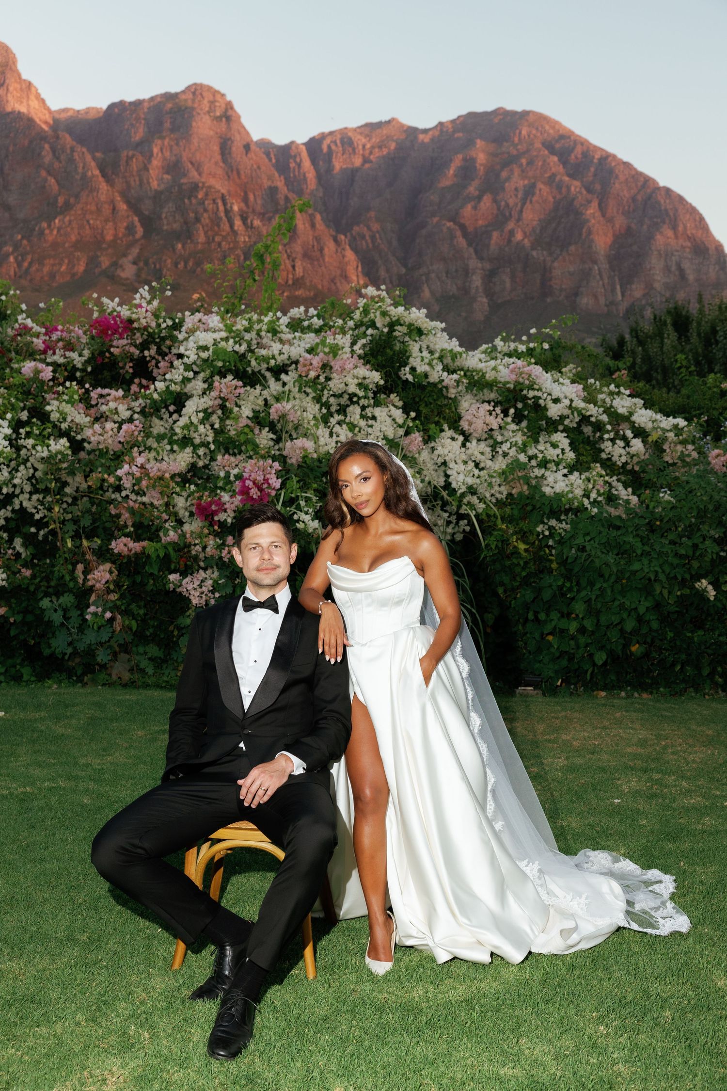 A wedding couple poses on grass with red rock mountains and flowering bushes in the background at sunset.